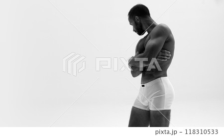 Side profile of man in white boxer briefs, his gaze down and arms crossed against white studio background. Monochrome filter. Side profile of man in white boxer briefs, his gaze down and arms crossed against white studio background. Monochrome filter. 118310533
