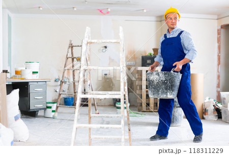 Worker in blue overalls and hardhat dragging various painting tools in room being renovated Worker in blue overalls and hardhat dragging various painting tools in room being renovated 118311229
