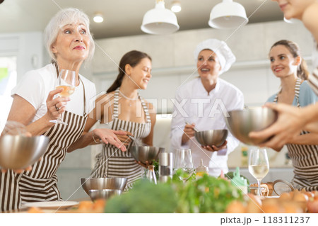 Aged woman holding wine and talking to other cooking course members whipping souce in bowls 118311237
