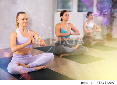 Sporty young women sitting on mats in Lotus Position Padmasana during yoga stretch joint workout in fitness center Sporty young women sitting on mats in Lotus Position Padmasana during yoga stretch joint workout in fitness center 118311289