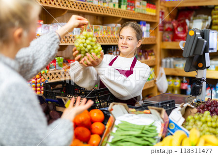 Girl who works part-time as a trainee saleswoman handing grape to customer after payment at checkout Girl who works part-time as a trainee saleswoman handing grape to customer after payment at checkout 118311477