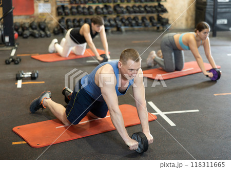 Focused middle-aged sportsman exercising with abs roller on fitness mat during group training in CrossFit center 118311665