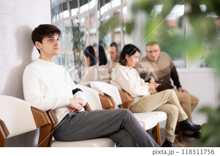 Young man browsing websites on smartphone while sitting in lobby of job center or employment office, waiting for interview or conversation with career counselor Young man browsing websites on smartphone while sitting in lobby of job center or employment office, waiting for interview or conversation with career counselor 118311756