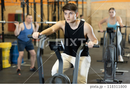 Energetic young male athlete training with exercise bike machine near other people gym 118311800