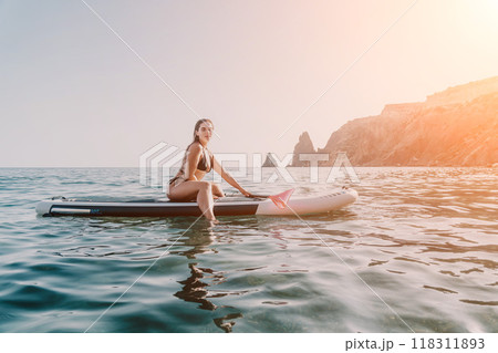 Woman Paddleboarding Ocean Coastline Summer - A woman in a bikini sits on a paddleboard in the ocean, with a mountainous coastline in the background. The sun is shining and the water is calm. This is Woman Paddleboarding Ocean Coastline Summer - A woman in a bikini sits on a paddleboard in the ocean, with a mountainous coastline in the background. The sun is shining and the water is calm. This is 118311893