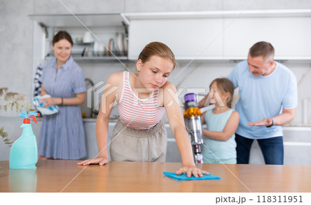 Bored teenage girl wiping table while cleaning apartment with family 118311951