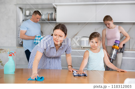 Cheerful woman wiping table in kitchen with preteen daughter Cheerful woman wiping table in kitchen with preteen daughter 118312003
