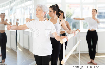 Elderly woman practicing ballet moves at barre during group class Elderly woman practicing ballet moves at barre during group class 118312219
