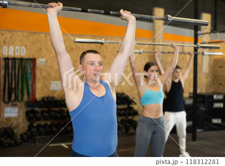 Motivated man practicing exercises with rod near other people in gym Motivated man practicing exercises with rod near other people in gym 118312281