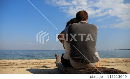 Romantic couple hugs during sitting on wooden log on the beach. Happy pair admiring a sea and landscape. Lovers spending time together at resort. Concept of vacation or holiday. Slow motion Romantic couple hugs during sitting on wooden log on the beach. Happy pair admiring a sea and landscape. Lovers spending time together at resort. Concept of vacation or holiday. Slow motion 118313649