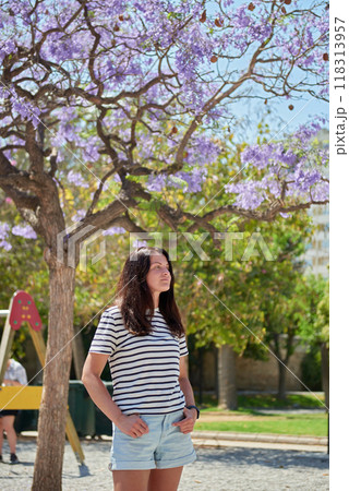 Woman in denim shorts near blooming tree, child swinging behind, warm sunny day, tourism, family holiday 118313957