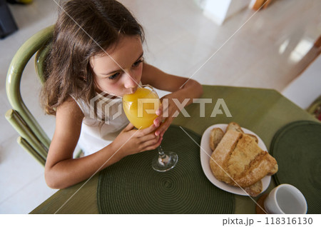 A young girl drinks orange juice at a table set with bread and a cup, exuding a refreshing breakfast vibe. A young girl drinks orange juice at a table set with bread and a cup, exuding a refreshing breakfast vibe. 118316130