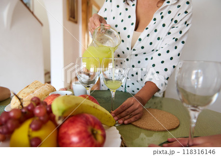 A woman pours fresh juice into glasses on a table adorned with fruit and bread, creating a serene and inviting kitchen scene. A woman pours fresh juice into glasses on a table adorned with fruit and bread, creating a serene and inviting kitchen scene. 118316146