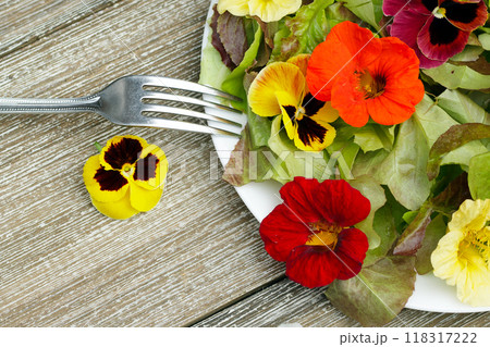 Table top view of the plate with salad made of edible flowers (nasturtium, pansy) and lettuce on the wooden table with a fork. 118317222