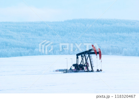 Beautiful winter landscape in prairies. Pumpjack in the middle of agricultural white field and the forest on hills covered with snow. Beautiful winter landscape in prairies. Pumpjack in the middle of agricultural white field and the forest on hills covered with snow. 118320467