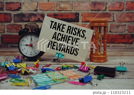 Take risks, achieve rewards text on paper card with alarm clock and stationary on wooden background 118323211