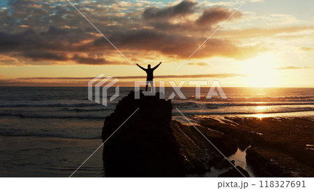 Human with raised hands looks to the sun over horizon in morning while sunrise. Man stands on a rock against the ocean beautiful summer sunset. Beauty of nature. Silhouette of a man against the sky Human with raised hands looks to the sun over horizon in morning while sunrise. Man stands on a rock against the ocean beautiful summer sunset. Beauty of nature. Silhouette of a man against the sky 118327691