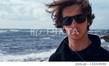 Young man with blown brown hair against the ocean on summer sunny day. Attractive young man looks at the camera on the beach. Handsome guy in black sunglasses poses outdoors on vacation. 118328098