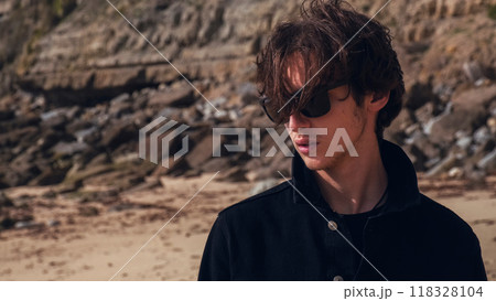Portrait of an young man with brown hair blown on wind against rock on summer sunny day. Attractive young man looks at the camera on the nature. Handsome guy in black sunglasses poses outdoors. 118328104