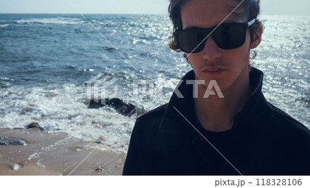 Young man with blown brown hair against the ocean on summer sunny day. Attractive young man looks at the camera on the beach. Handsome guy in black sunglasses poses outdoors on vacation. 118328106