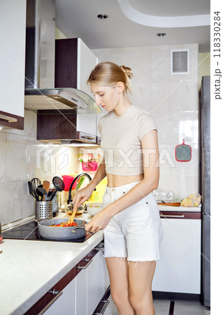 Young woman cooking in a modern kitchen with fresh vegetables in a frying pan during the day Young woman cooking in a modern kitchen with fresh vegetables in a frying pan during the day 118330284