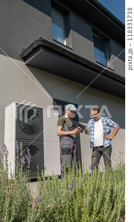 A technician and a homeowner discussing heat pump maintenance outside a house, with a heat pump unit visible in the background. A technician and a homeowner discussing heat pump maintenance outside a house, with a heat pump unit visible in the background. 118331738