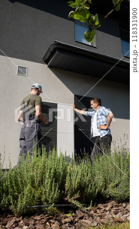 A technician and a homeowner inspecting a heat pump unit outside a house in the garden. 118331900