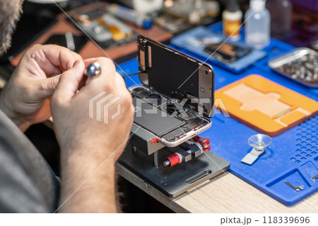 A skilled technician performs phone repair and replaces a battery in a well-organized workshop with tools and parts readily available. 118339696