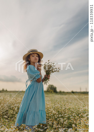 Brooding natural girl in blue linen dress and canotier straw hat stands and holds bouquet of flowers in daisy field, vintage photo Brooding natural girl in blue linen dress and canotier straw hat stands and holds bouquet of flowers in daisy field, vintage photo 118339931