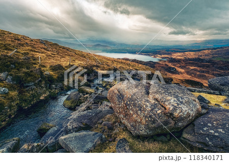 A breathtaking view of rocky terrain leads down to a serene lake, with rolling hills in the background under a moody sky filled with clouds. 118340171