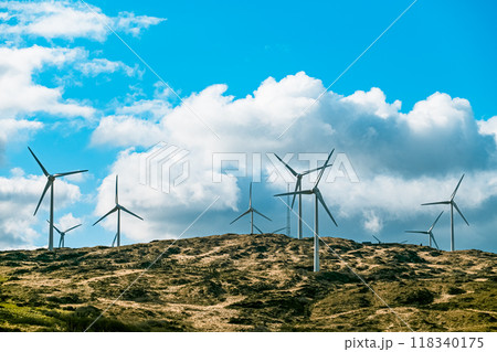 Wind turbines are seen on a rugged hillside, with a partly cloudy sky above. 118340175