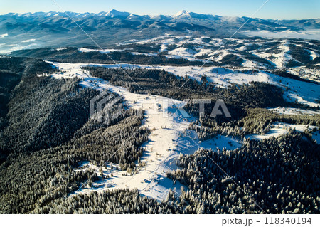 Aerial view of snow-covered valley with winding road passing through small village. Landscape features gently rolling hills, dense forests, and distant mountains under clear blue sky, with soft mist. Aerial view of snow-covered valley with winding road passing through small village. Landscape features gently rolling hills, dense forests, and distant mountains under clear blue sky, with soft mist. 118340194