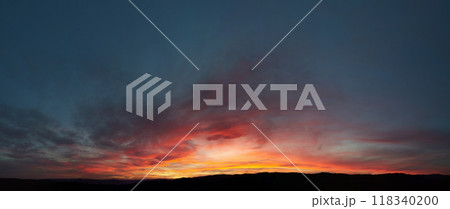 Stunning sunset paints sky with vibrant hues of red, orange, and pink, blending into deep blues and purples. Silhouetted hills create contrast against fiery horizon, adding drama to serene landscape. 118340200