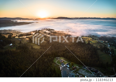 Aerial view of misty morning in mountains, with small village nestled among trees. Fog gently blankets landscape, creating serene and picturesque scene, as sun begins to rise in the background. 118340213