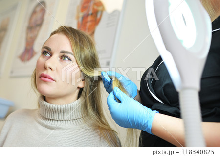 A dermatologist trichologist examines the hair structure of a young woman's patient using an optical dermatoscope device. 118340825