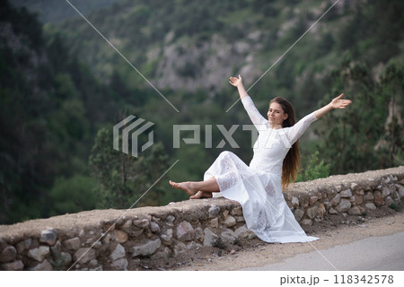 A woman in a white dress is sitting on a rock by a road. She is smiling and she is happy. 118342578