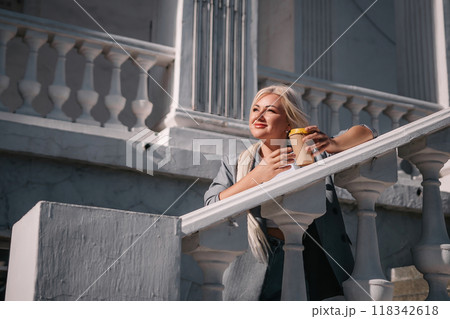 A woman is standing on a white railing with a cup of coffee in her hand. She is wearing a gray jacket and blue jeans. Concept of relaxation and leisure. 118342618