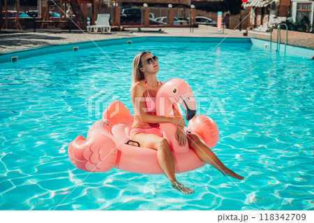 A woman is sitting on a pink inflatable flamingo in a pool. Concept of fun and relaxation, as the woman is enjoying her time in the water 118342709