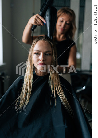 Vertical shot of female hairdresser drying and straightening long blonde customer hair with comb and hairdryer in luxury beauty salon. Attractive lady client getting stylish hairdo done at salon. Vertical shot of female hairdresser drying and straightening long blonde customer hair with comb and hairdryer in luxury beauty salon. Attractive lady client getting stylish hairdo done at salon. 118343918
