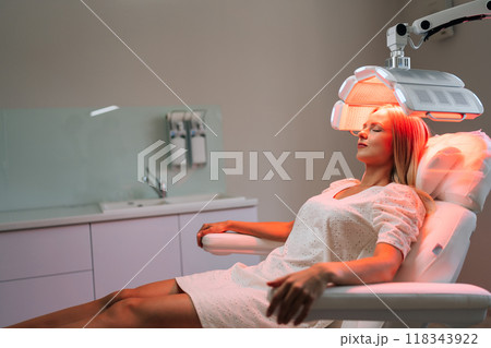 Side view of young woman with blond hair in white uniform having LED light hair photodynamic therapy during hair treatment at medical clinic. Female client having non-invasive type of phototherapy. Side view of young woman with blond hair in white uniform having LED light hair photodynamic therapy during hair treatment at medical clinic. Female client having non-invasive type of phototherapy. 118343922