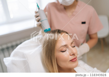 Closeup face of attractive blonde female patient having hair light therapy with modern innovation apparatus. Trichologist performing hair treatment procedure with spectrum phototherapy device. Closeup face of attractive blonde female patient having hair light therapy with modern innovation apparatus. Trichologist performing hair treatment procedure with spectrum phototherapy device. 118343928