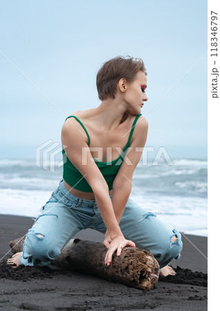 Woman is having blast in playful pose on log, sitting like she's riding horse and turning head to side with those flirty eyes closed. Woman in jeans and top is just chilling on summer beach 118346797