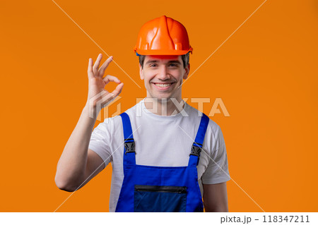 Positive builder in helmet, protective uniform makes OK okay hand sign on orange Positive builder in helmet, protective uniform makes OK okay hand sign on orange 118347211