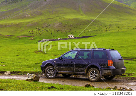 black car on a side of wet dirt road in the mountain hills in front of sheep grazing on green pasture at rainy day near farm ruins 118348202