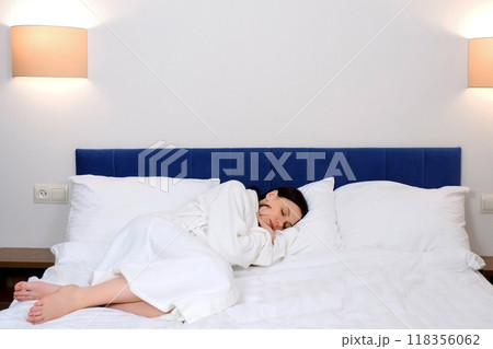 Tired young brunette woman in white bathrobe is sleeping in bed in hotel alone. Tired young brunette woman in white bathrobe is sleeping in bed in hotel alone. 118356062