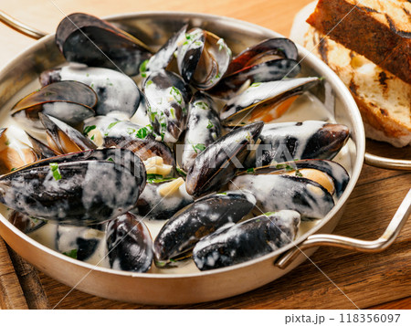 Delicious black mussels in creamy cheese and garlic sauce. Mediterranean black mussels in metal pan on wooden cutting board with grilled bread on wooden background Delicious black mussels in creamy cheese and garlic sauce. Mediterranean black mussels in metal pan on wooden cutting board with grilled bread on wooden background 118356097