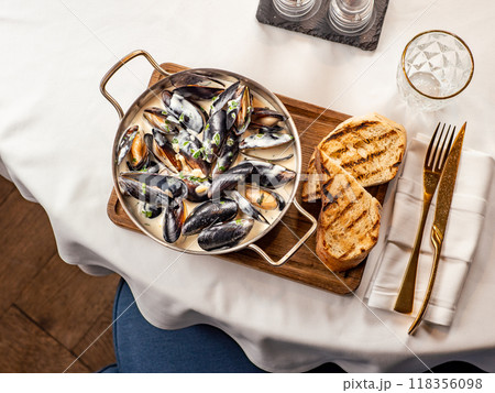 Delicious black mussels in creamy cheese and garlic sauce. Mediterranean black mussels in metal pan on wooden cutting board with grilled bread on restaurant table background, top view 118356098