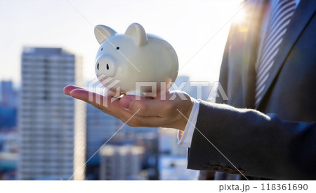 ブタの貯金箱を手の上に乗せたスーツを着た男性。貯金や預金額のイメージ。都市の高層ビルの背景。 118361690
