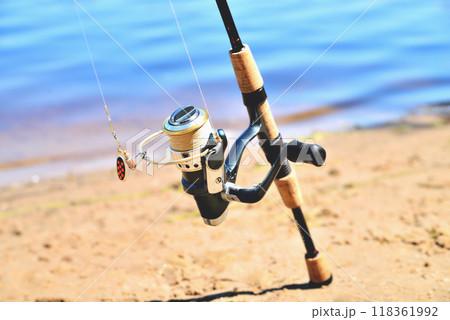 fishing rod with reel on the background of the river, blue water, sun, sand fishing rod with reel on the background of the river, blue water, sun, sand 118361992