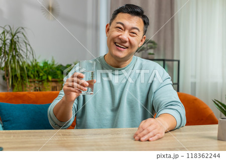 Thirsty Asian man sits at home table holding glass of natural aqua make sips drinking filtered water 118362244